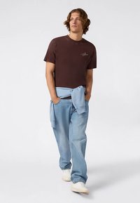 Bruine katoenen T-shirt met ronde hals, gecombineerd met lichtblauwe spijkerbroek. Een lichtblauwe trui om de taille geknoopt. Witte sneakers.