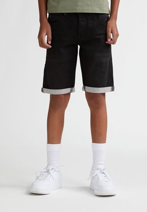 Shorts en denim noir avec ourlet retroussé, présentant un revers gris. Associés à des baskets blanches et des chaussettes hautes, posés sur un fond uni.
