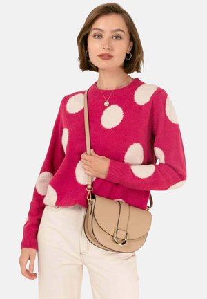 Femme portant un pull rose à gros pois blancs, un pantalon crème et un sac bandoulière beige avec une boucle dorée.