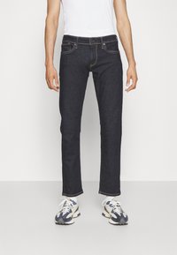 Mörk denimjeans med rak passform, femfickorsdesign, kontrasterande sömmar och knappstängning, kombinerat med vit t-shirt och sneakers.