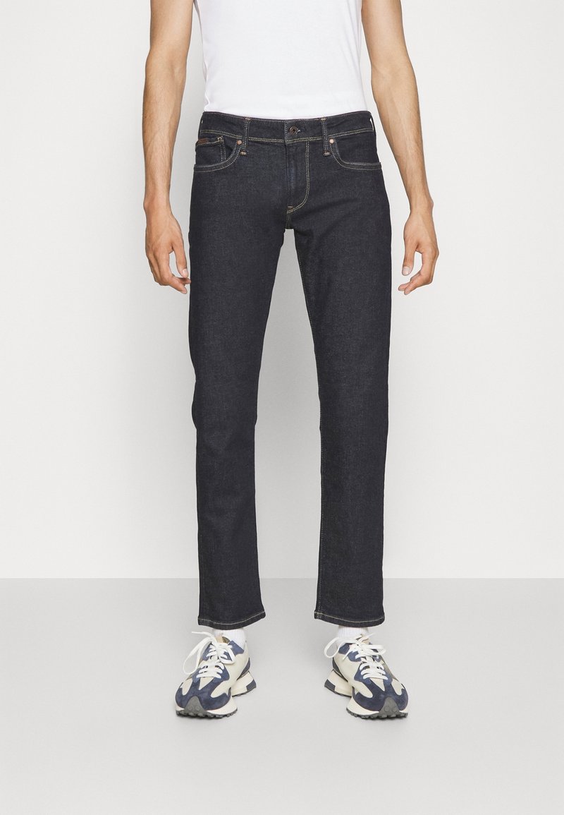 Mörk denimjeans med rak passform, femfickorsdesign, kontrasterande sömmar och knappstängning, kombinerat med vit t-shirt och sneakers.