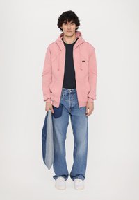 Helles rosa Kapuzensweatshirt mit Kordeln, kombiniert mit einem dunkelblauen T-Shirt und lockeren Jeans in Denim. Weiße Sneakers und eine graue Jacke.