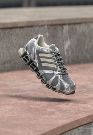 Pantof sport gri și argintiu cu talpă unică asemănătoare unui arc, plutind deasupra unor trepte din beton rozalii.