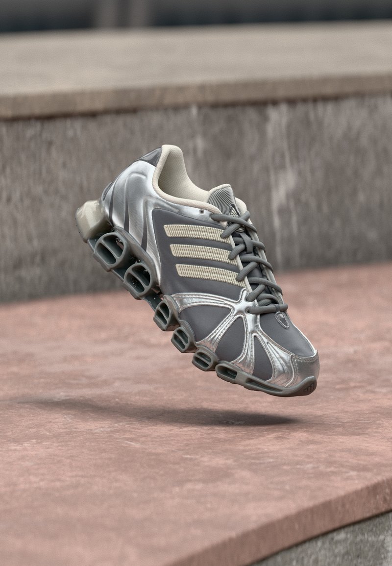 Γκρι και ασημί αθλητικό παπούτσι με μοναδική σόλα σε στυλ ελατηρίου που αιωρείται πάνω από ροζ σκουρόχρωμα σκάλες από σκυρόδεμα.