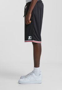 Pantalones cortos deportivos negros con acentos rojos y blancos, con un logo en la pierna izquierda. Material ligero, cinturilla elástica y bolsillos laterales.