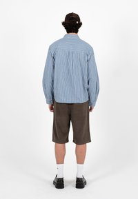 Jeune homme debout de dos, portant une chemise à manches longues à rayures bleues et blanches, un short brun jusqu'aux genoux, des chaussures noires et une casquette sombre.