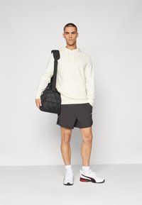 Hoodie beige chiaro con tasca frontale, borsa nera regolabile, pantaloni corti grigio scuro, scarpe sportive bianche con dettagli rossi, in piedi contro uno sfondo neutro.