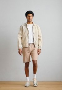 Μπεζ μπουφάν μπόμπερ πάνω από λευκό t-shirt, ανοιχτό καφέ σορτς, λευκές κάλτσες, αθλητικά παπούτσια με πράσινες λεπτομέρειες, στέκεται μπροστά σε ουδέτερο φόντο.