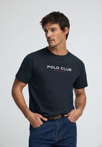 Mörk marinblå T-shirt med korta ärmar som har en vit och röd "POLO CLUB"-logotyp, kombinerad med blå jeans och ett brunt bälte.