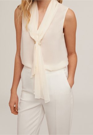 Vrouw draagt een mouwloze crèmekleurige blouse met sjaal die vastgeknoopt is met een pareldetail, gecombineerd met witte hoge taillebroek, hand in de zak.
