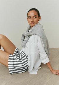 Camicia bianca abbottonata, felpa grigia drappeggiata sulle spalle e pantaloncini a righe grigie e bianche. Tessuto liscio, design casual, posa seduta.