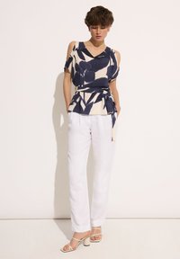 Bluzka w kwiatowy wzór w odcieniach kremu i granatu, z wycięciami na ramionach. Połączona z białymi lnianymi spodniami oraz beżowymi sandałami na słupku.