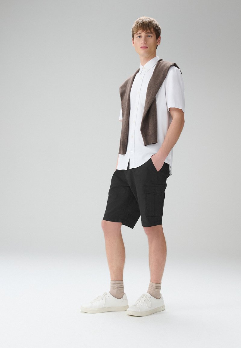 Joven de pie con las manos en los bolsillos, lleva una camisa blanca de manga corta, pantalones cortos negros, un suéter beige sobre los hombros, calcetines beige y zapatillas blancas.