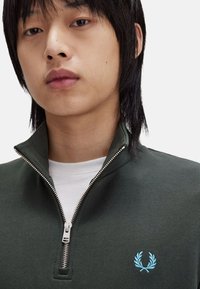 Suéter verde con cremallera y cuello alto, con cremallera plateada y un logotipo bordado en azul claro en el lado izquierdo del pecho. La capa interior es blanca.