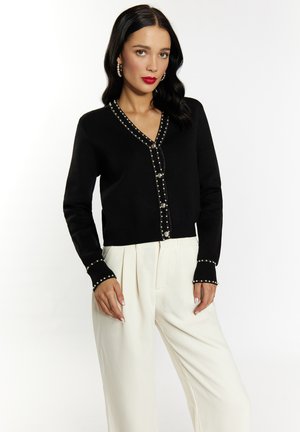 Schwarze kurz geschnittene Strickjacke mit V-Ausschnitt, verzierten Knöpfen und Perlenakzenten an den Ärmeln. Kombiniert mit weißen, plissierten Hosen.