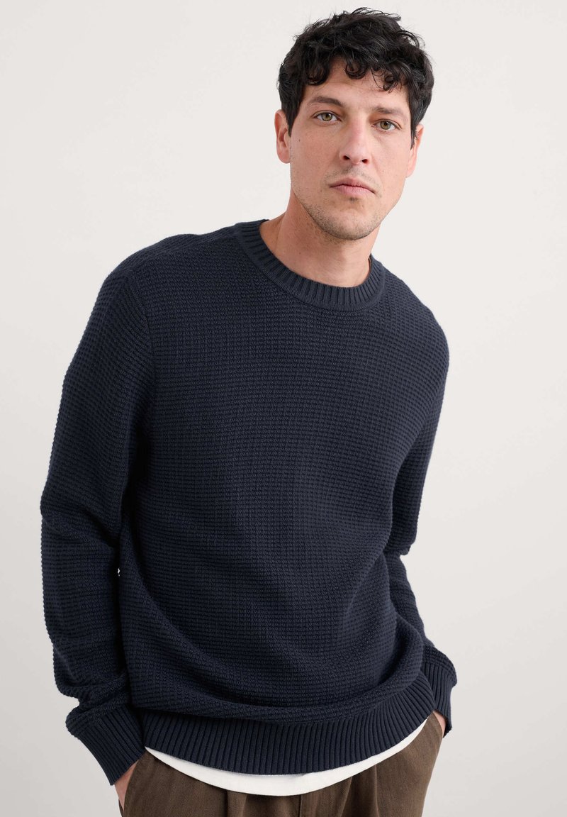 Mörkblå texturerad tröja med rund halsringning, ribbade ärmmuddar och nederkant. Bärs över en vit skjorta med avslappnad passform och subtilt mönster.