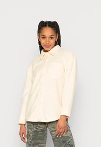 Camisa de fleece bege claro com botões e colarinho, um bolso no peito e detalhes de costura visíveis. Combinada com calças de camuflagem.