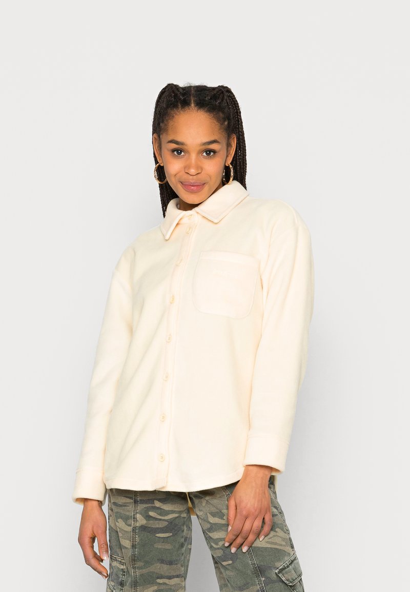 Camisa de fleece bege claro com botões e colarinho, um bolso no peito e detalhes de costura visíveis. Combinada com calças de camuflagem.