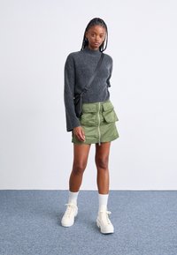 Pull en tricot gris, jupe zippée verte avec poches cargo, chaussettes blanches et baskets à plateforme beiges. Sac bandoulière simple comme accessoire.