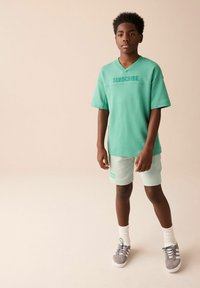 Mintgrøn oversized t-shirt med "SUBSCRIBE" trykt i mørkegrøn, parret med lyse mintfarvede shorts. Grå sneakers og hvide sokker fuldender looket.