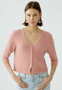 Hellrosa, verkürzter Cardigan mit V-Ausschnitt, kurzen Ärmeln und fünf sichtbaren Knöpfen. Weicher Stoff und gerippter Saum. Über leichter Jeans getragen.