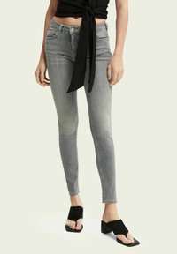 Jeans skinny grises con un acabado desgastado y ligera distorsión, combinados con tacones abiertos negros y una blusa negra anudada.