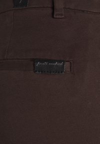 Primo piano di un tessuto marrone scuro di pantaloni con un piccolo etichetta in pelle nera cucita sopra una tasca, che presenta un testo cursivo bianco.