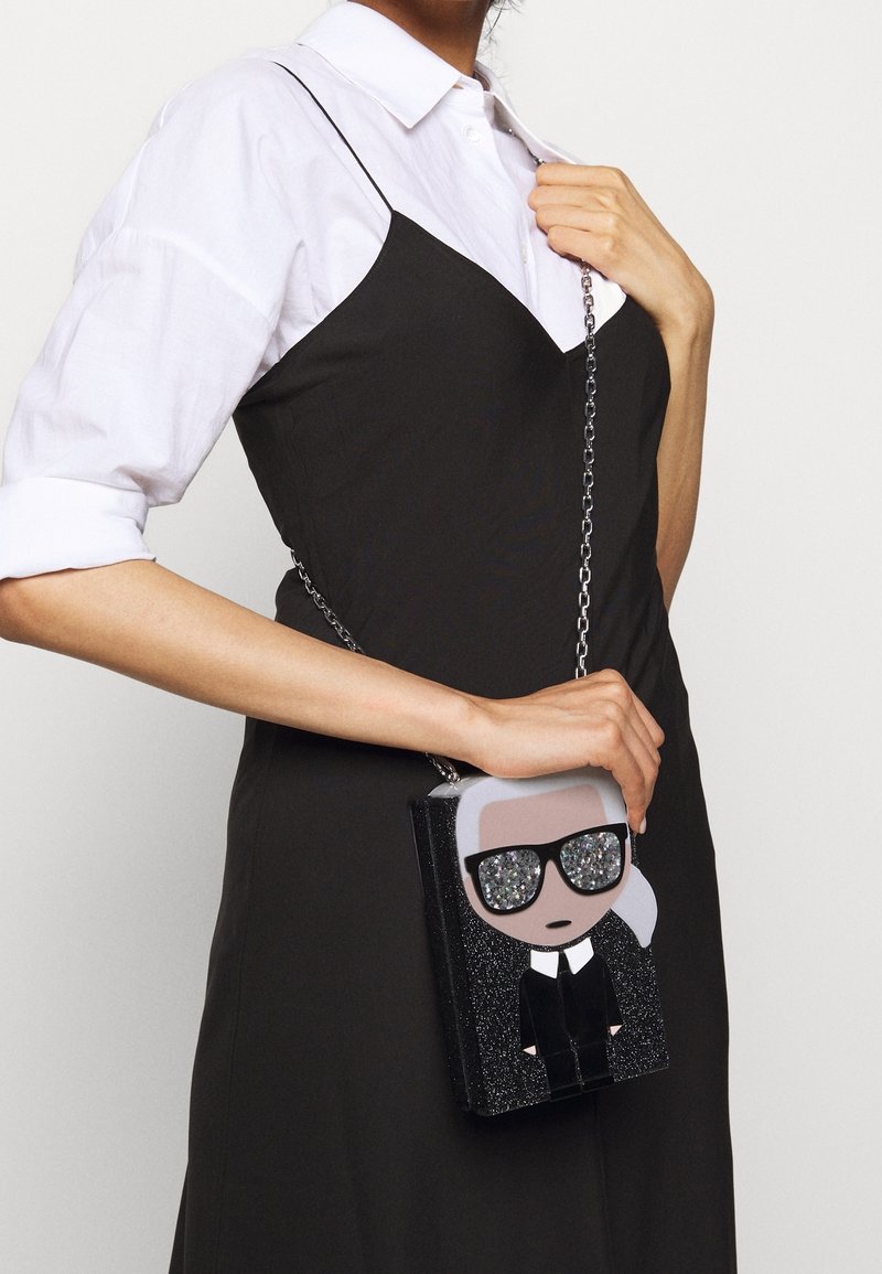 Czarna torebka błyszcząca, w kształcie postaci z srebnymi włosami i okularami przeciwsłonecznymi; ma srebrny łańcuszek i błyszczące detale.
