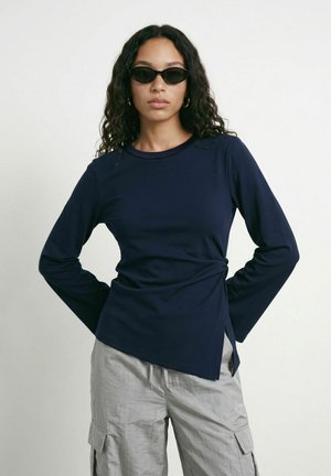 Femme aux cheveux bouclés portant des lunettes de soleil noires, un haut à manches longues bleu marine et un pantalon cargo gris clair, debout les mains derrière le dos.