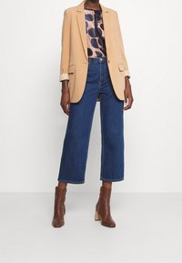 Beżowa marynarka z podwiniętymi rękawami noszona na wzorzystej bluzce, w połączeniu z szerokimi niebieskimi dżinsami i brązowymi botkami na obcasie.