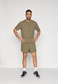 Αθλητικό πουκάμισο και σορτς σε ελιά πράσινο χρώμα, από ύφασμα, με μαύρο λογότυπο. Φορεμένα με μαύρα παπούτσια και λευκές κάλτσες αστραγάλου. Χωρίς συντροφιά, σε όρθια στάση.