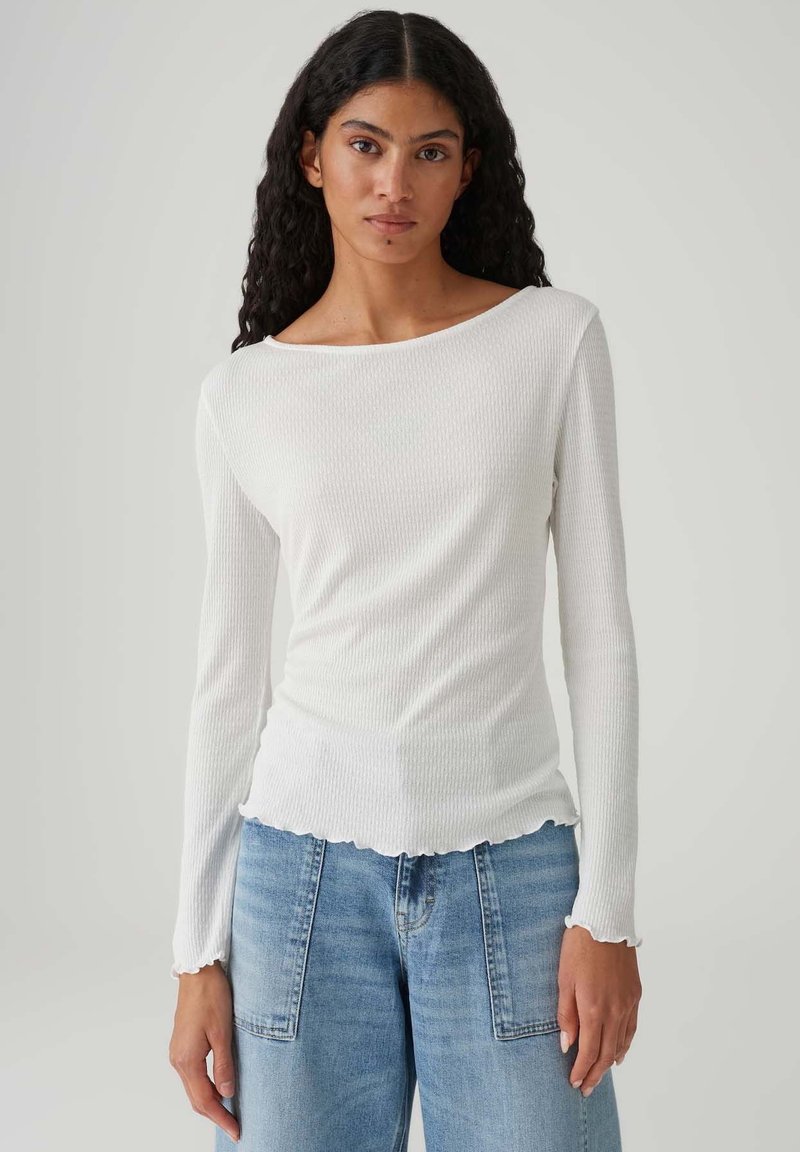 Weißes, langärmeliges, geripptes Oberteil mit Bootsausschnitt und wellenförmigem Saum. Kombiniert mit hellblauen Jeans mit weitem Bein. Texturiertes Stoffmaterial.