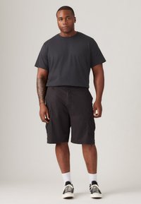 Svart t-skjorte med kort erme kombinert med svarte cargo-shorts. Antrekket har flere sidelommer, og det er kombinert med hvite sokker og svarte joggesko.