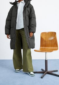 Zwarte pufferjas met een capuchon, wit gestreept shirt, olijfgroene wijde broek en casual groene en witte sneakers naast een houten stoel.