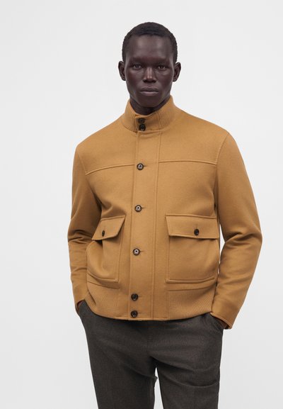 Homme portant une veste beige boutonnée avec deux poches avant à rabat et un pantalon gris foncé, debout, mains dans les poches, devant un fond uni.
