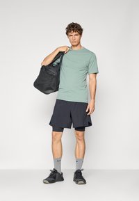 Camiseta de manga corta verde menta, pantalones cortos deportivos negros, calcetines grises y zapatillas negras. Llevando una bolsa de gimnasio negra con superficie texturizada.