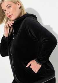 Sudadera con cremallera de terciopelo negro, con un cuello alto, bolsillos laterales y ribetes blancos en los puños. Textura suave y diseño ajustado.