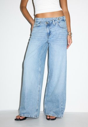 Femme portant un jean bleu clair à jambes larges avec un détail à double bouton, des sandales noires, un crop top blanc et un bracelet argenté au poignet droit.