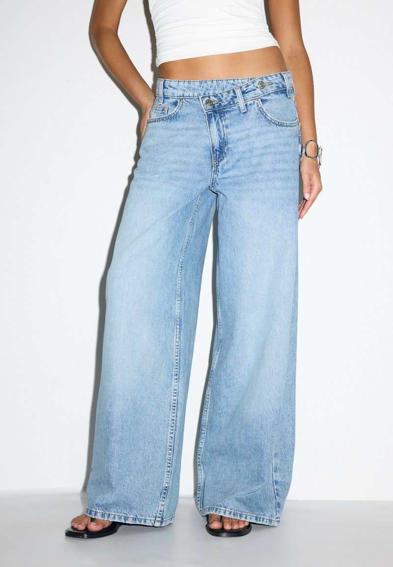 Femme portant un jean bleu clair à jambes larges avec un détail à double bouton, des sandales noires, un crop top blanc et un bracelet argenté au poignet droit.