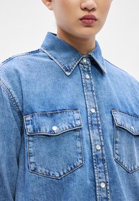 Modrá rifľová košeľa s golierom, zapínaním na gombíky, dvoma náprsnými vreckami a gombíkmi pripomínajúcimi perly. Má klasický strih a jemne vyblednutú textúru.