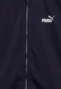 Tengerészkék széldzseki elülső cipzárral és fehér Puma logóval, sima felülettel és könnyű anyagból készült.