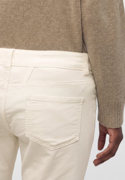 Personne portant un pull en maille beige et un pantalon blanc, montrant la poche arrière et le côté avec la main détendue sur le côté.