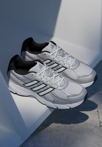 Biele a strieborné atletické tenisky s textíliou z sieťoviny, čiernymi akcentmi a šedými detailmi. Obsahujú tradičné šnúrky a textúrovanú podrážku.