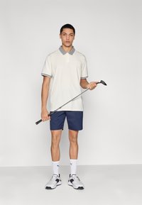 Φωτεινό μπεζ πόλο με γκρι γιακά, ναυτικά σορτς, κρατώντας ένα γκολφ κλαμπ. Άσπρες κάλτσες και αθλητικά παπούτσια ολοκληρώνουν το σύνολο.