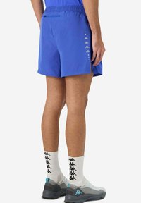 Pantaloni sportivi blu con vita regolabile tramite cordino, tasche laterali e logo Kappa bianco. Indossati con scarpe grigie e calze bianche con logo nero.