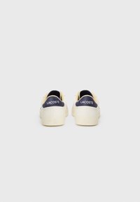 White sneakers with a smooth texture, featuring a navy heel tab with "LACOSTE" logo, and a minimalist design. Rubber sole, lace-up closure.