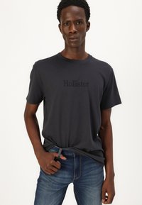 T-shirt gris foncé à manches courtes avec le logo "Hollister" en lettres en relief, associé à un jean en denim bleu avec une texture subtile.