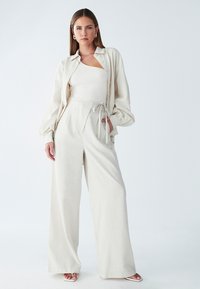 Kremno obarvana lanena jakna preko rebraste bele bodi majice, v kombinaciji z visokim pasom in širokimi hlačami ter belimi sandalami s trakovi.