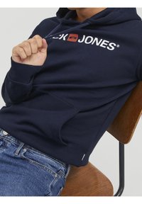 Tmavě modrá bavlněná mikina s kapucí, vybavená přední kapsou, šňůrkami na stažení a bílým a červeným logem přes hruď. Viditelné světlé modré džíny.