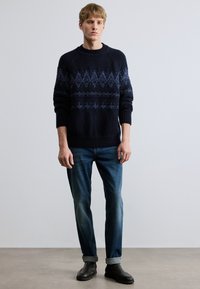 Tengerészkék kötött pulóver bonyolult kék mintázattal, kerek nyakkivágással és bordázott mandzsettákkal, sötétkék farmernadrággal és fekete bokacsizmával párosítva.